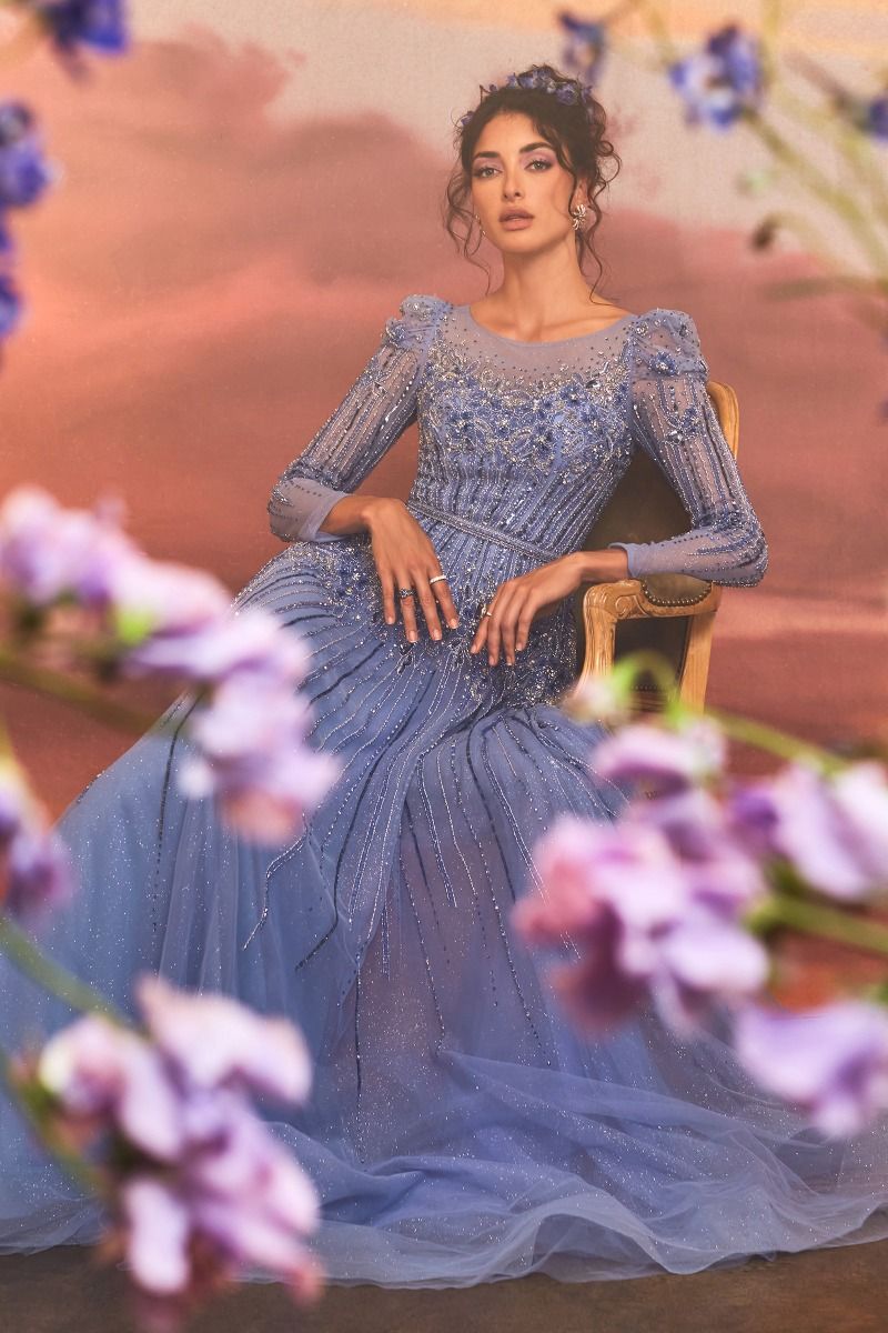 A woman in the Luxurious Formal Gown A1167 by Andrea and Leo sits on a gold chair, with blurred purple flowers around her against a warm background.
